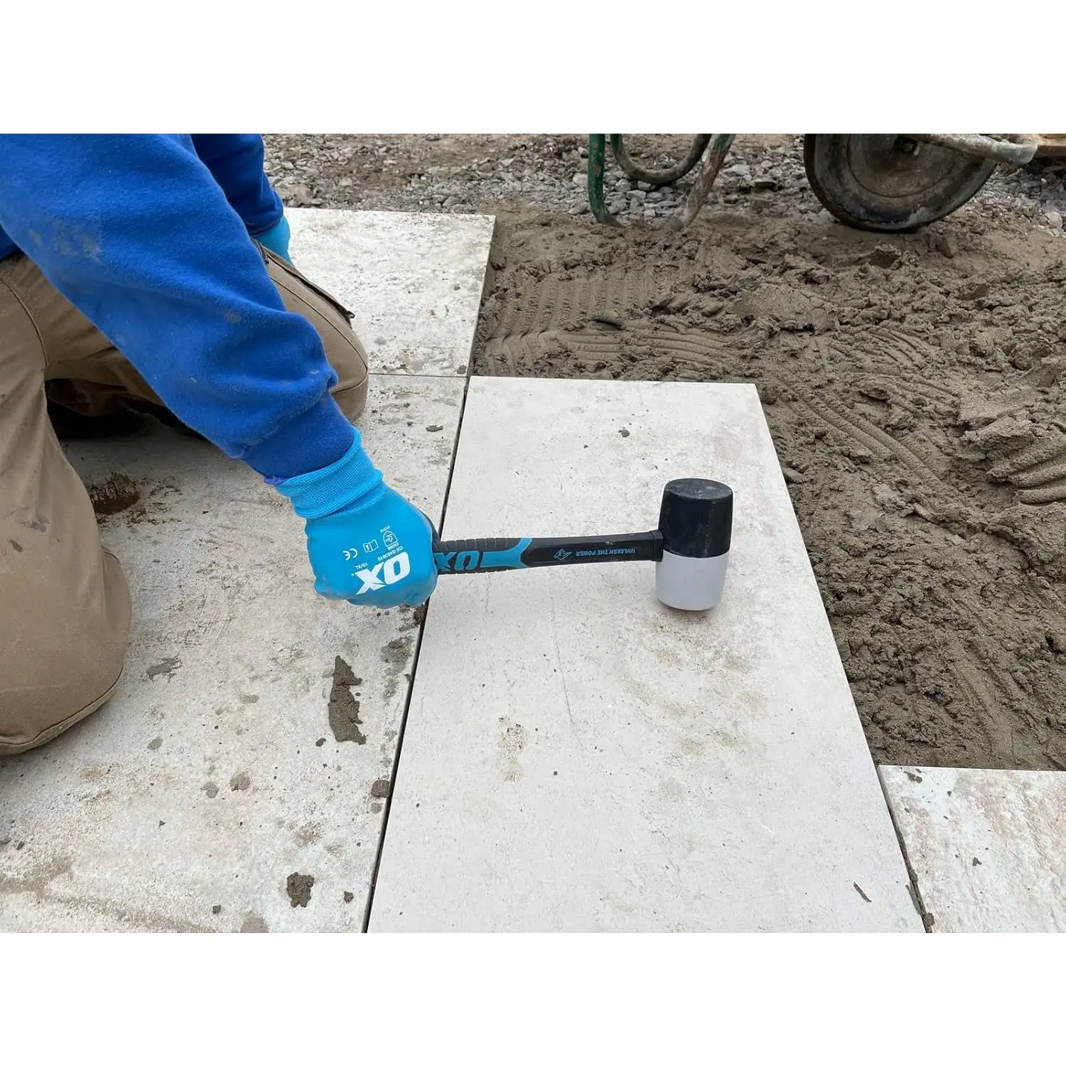 Persoană cu mănuși albastre și tricou albastru folosind ciocan cauciuc combinat OX Trade cu soft grip pentru a poziționa pavele